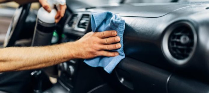 A person is holding a towel in one hand and a spray bottle in the other. He is wiping down the dashboard of a car.