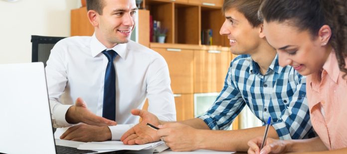 A couple sitting at a desk with a businessman. The woman signs some paperwork while the two men reach out to shake hands.