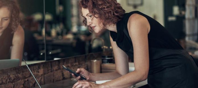 A woman hunched over at a table with her laptop, phone, and calculator out. She is standing next to a very large salon mirror.