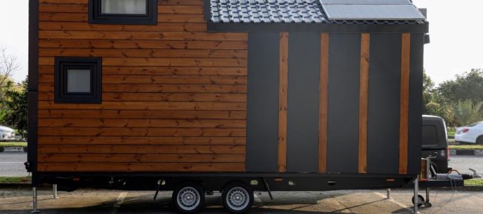 A tiny house attached to a trailer and in a parking lot. The home features wooden siding panels and small windows.