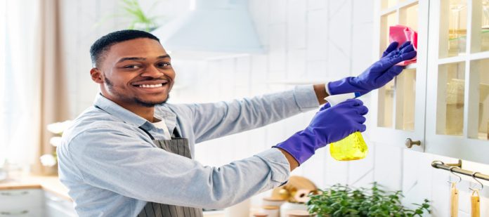 A man smiles as he cleans his kitchen's cabinets. He wears a green kitchen apron and purple kitchen gloves.