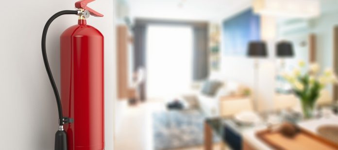 A close-up of a fire extinguisher mounted on a wall inside a home's kitchen, blurred in the background.