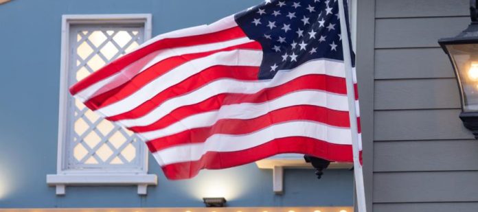 An American flag on a fiberglass flagpole ripples in the wind. A home with a white lattice window is in the background.