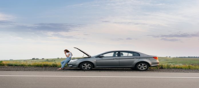 A frustrated young woman stands near a broken-down car with the hood up in the middle of the highway during sunset.