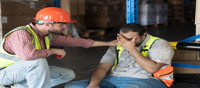 A warehouse employee struggles on the floor as another worker consoles them nearby. They wear safety vests.