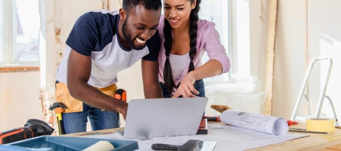 A man and woman at a workbench in the middle of a room under renovations. They have schematics and a laptop on the bench.