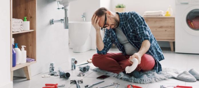 A man sitting on a bathroom floor with his head in his hand. He is looking at an array of tools spread out in front of him.