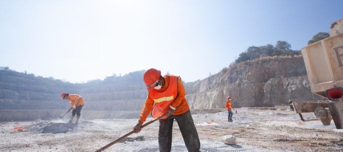 Three workers in high-vis orange safety gear stand in a dusty work site with rakes as the dust flies around them.