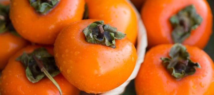 Five ripe persimmons piled in a small bowl sitting on a table. Three more persimmons sit beside the bowl.