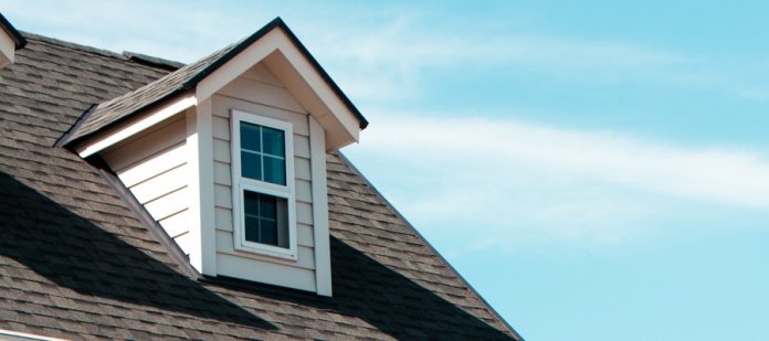 A low-angle view of a two-story house with a small dormer window protruding from the roof against a light blue sky.