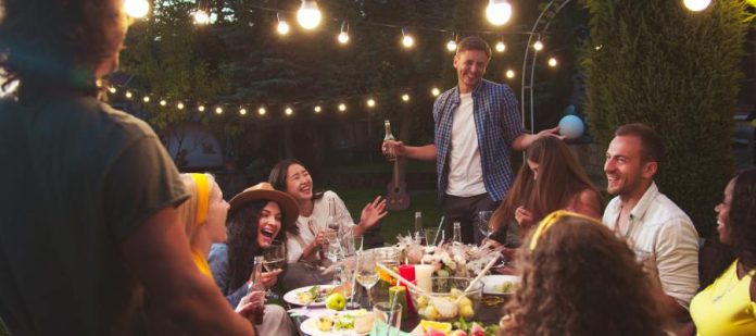 A group of people have fun and laugh while standing or sitting around a table at an outdoor backyard party.