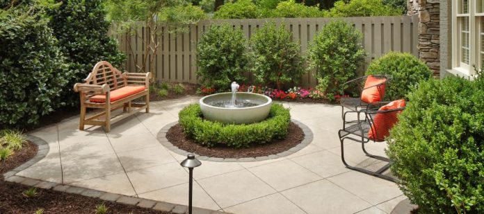 A concrete patio with a circular water fountain and outdoor furniture surrounded by a landscaped garden.