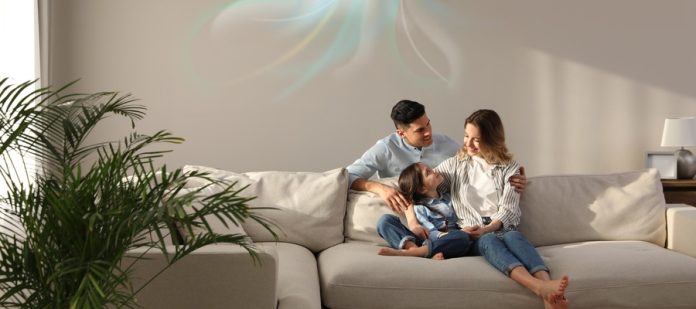 A family resting on the couch in their living room. There is an AC unit on the wall above them, keeping the house cool.