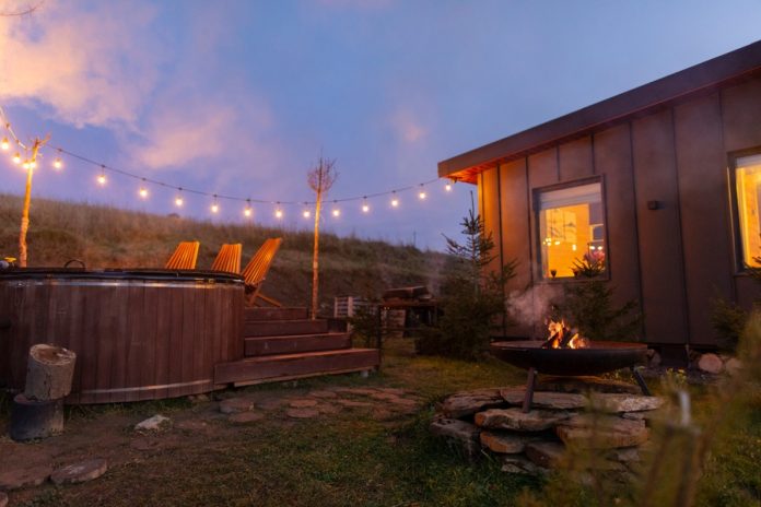 A backyard space with a seating area next to a hot tub, a fire pit on a stack of rocks, string lights, and a grill.