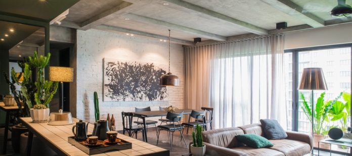 Modern apartment interior with a leather sofa, dining table, white brick wall art, sheer curtains, and greenery by windows.