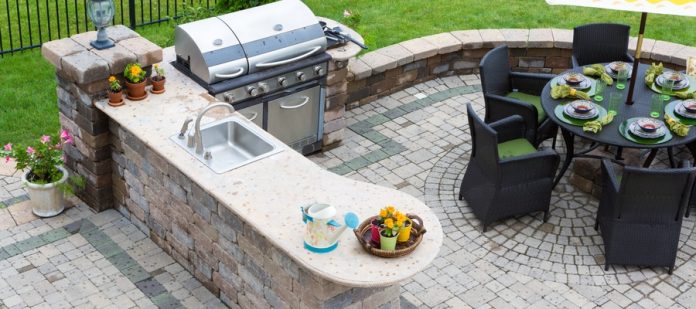 An outdoor kitchen with a built-in grill and sink on a stone patio beside a dining table with umbrella and chairs.