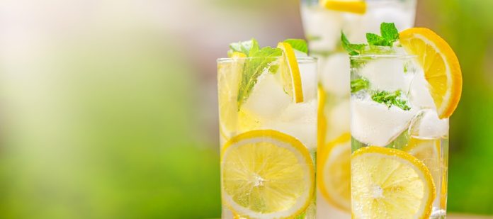 A sunny day with a close-up of three tall glasses of lemonade sitting on the table. The glasses also have mint sprigs.
