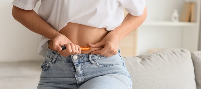 Someone stands with their shirt lifted, exposing their stomach. They hold an injectable medication up to their stomach.