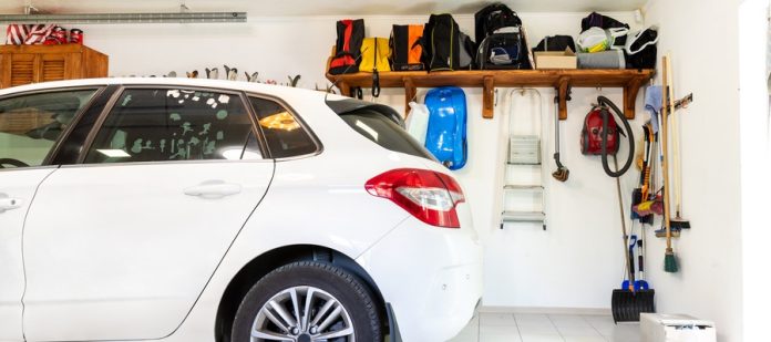 A white vehicle is parked in a garage with white tiled flooring. Bags, brooms, and lawn tools are on a wall-mounted shelf.