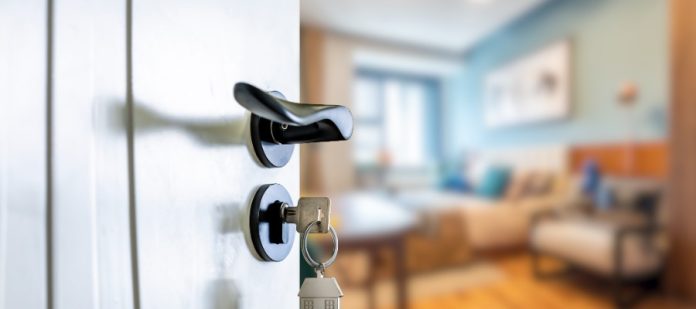A key rests in a white door lock with a black handle and house-shaped keychain beside a blurred room.