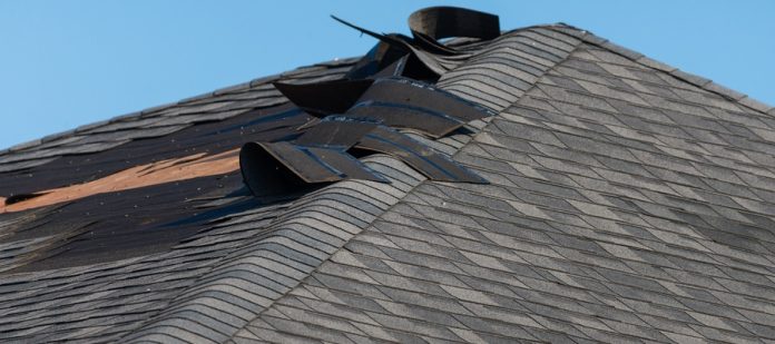 A storm-damaged asphalt shingle roof with torn and lifted shingles exposing underlayment beneath a clear blue sky.