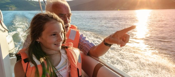 A girl and an older man wearing life jackets ride in a motorboat as the sun sets over a mountain lake.