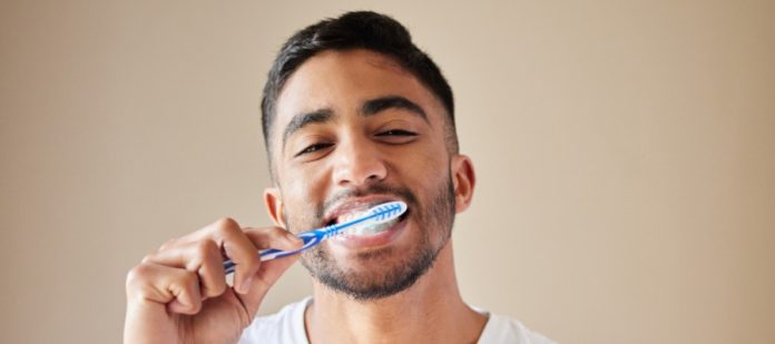 Smiling man in a white shirt brushes his teeth with a blue toothbrush against a plain beige background.
