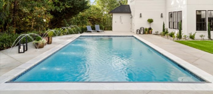 A rectangular backyard swimming pool with water fountains next to lounge chairs and a modern white two-story house.