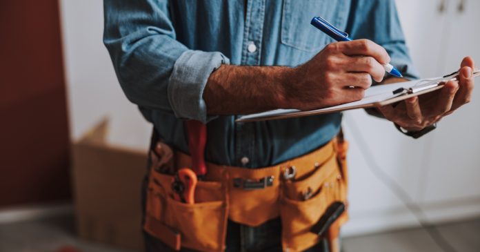 A person in a denim shirt writing on a clipboard while wearing a tool belt with hand tools near white cabinets.