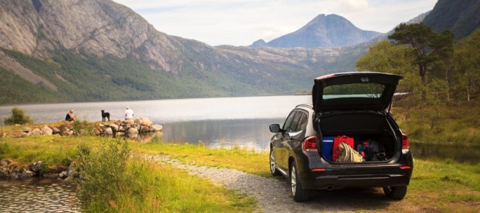 A car parked by a lake with its trunk open during the daytime. Two people and a dog are near the water.